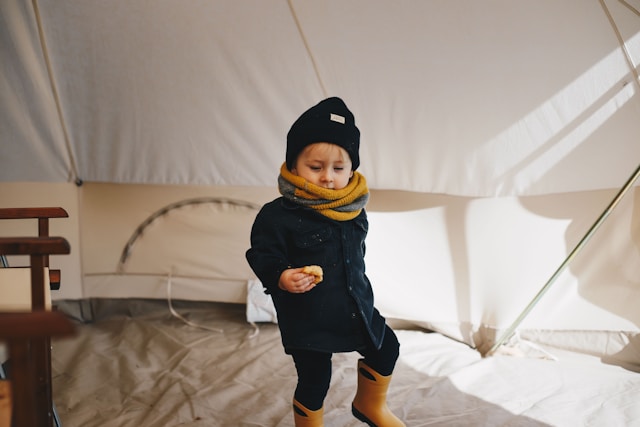 A small boy holding a piece of pastry in waterproof boots, a jacket, and a scarf, near a white tent.