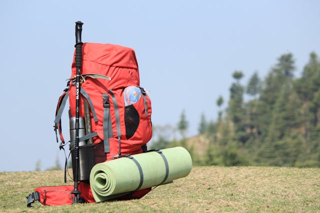 Red hiking backpack with attached trekking poles and a rolled sleeping mat on grass, showing how to protect outdoor equipment from moisture during outdoor trips